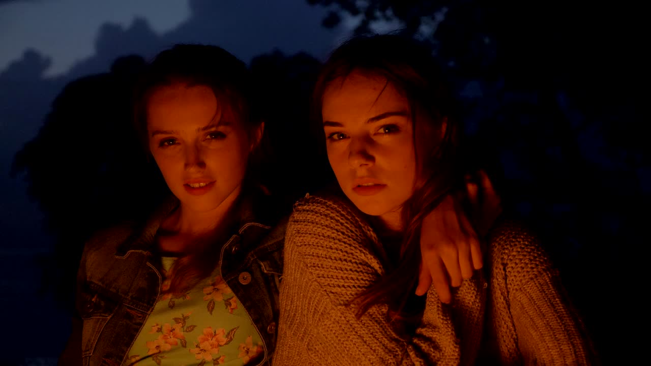 두 명의 젊은 백인 소녀가 저녁에 자연에서 불 에 앉아 카메라, 초상화를 바라보고 있습니다.