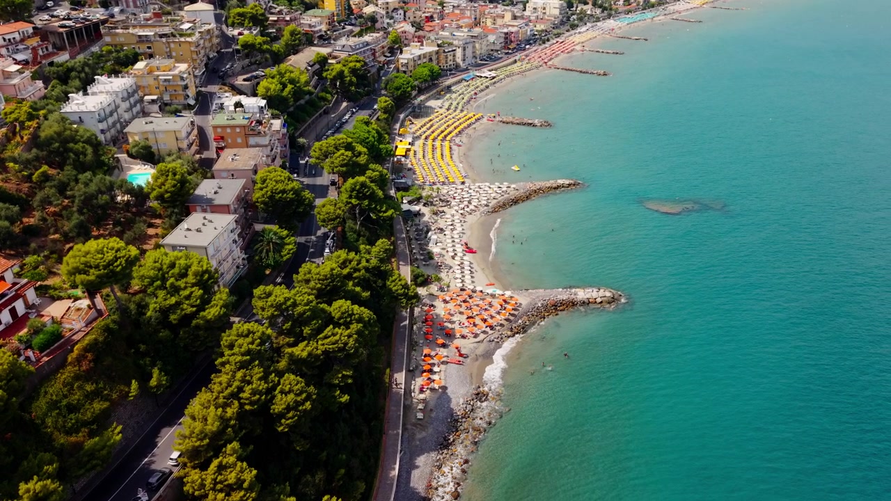 Tomada aérea de drone movendo-se diagonalmente em direção à costa de Laigueglia, mostrando praias organizadas, guarda-sóis coloridos, quebra-mares, estrada costeira e edifícios à beira-mar