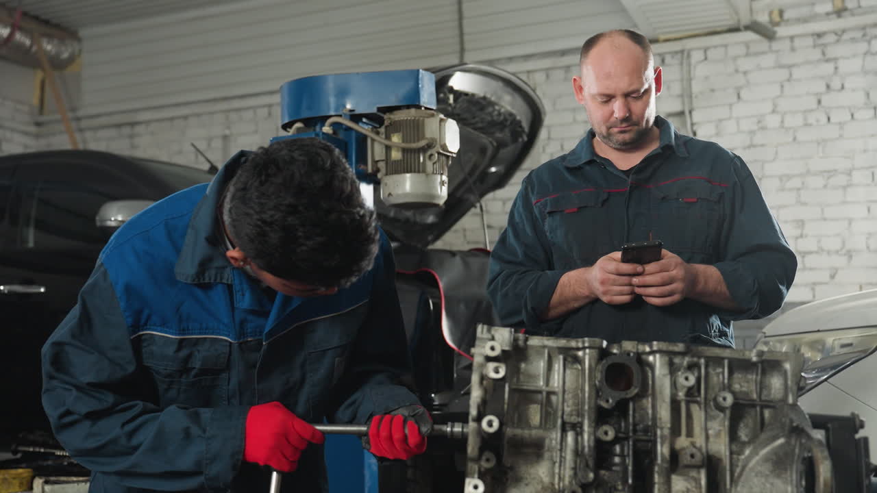 ingenieros mecánicos en un taller; uno trabajando atentamente en un motor con una llave inglesa mientras su colega usa su teléfono, iluminado por una luz brillante en el fondo