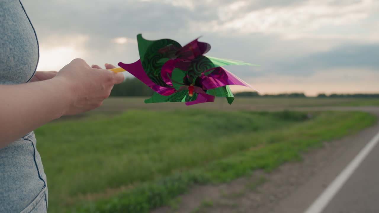 Close up rear view woman walking along rural road rotating pinwheel in hand with gentle breeze blowing through denim dress under overcast sky beside lush green field