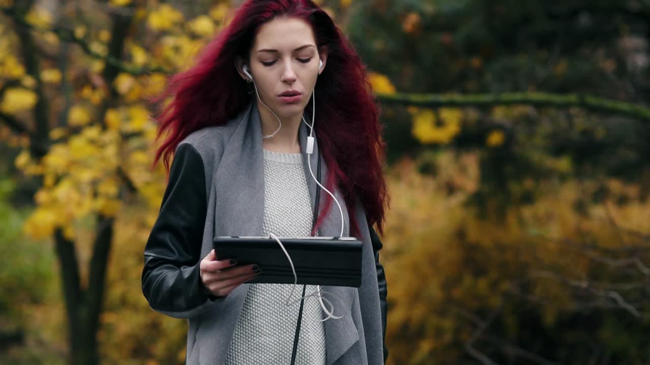 Young businesswoman with red hair walks in the autumn park and communicates via tablet pc. Young attractive woman with digital tablet computer in hands communicates using earphones