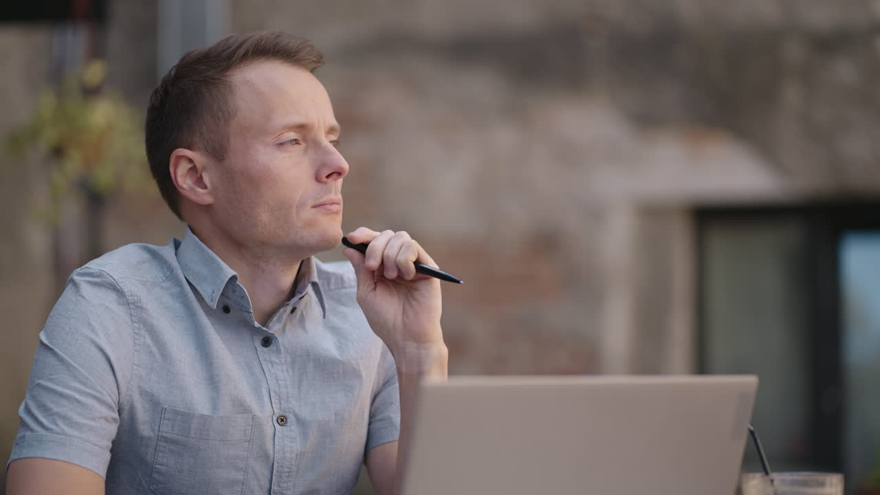 Close-up portrait of ponder young man in office. Designer plans his work and holding a pencil in his hand. Shooting is slow motion from below. Thoughtful serious man sit with laptop thinking solution