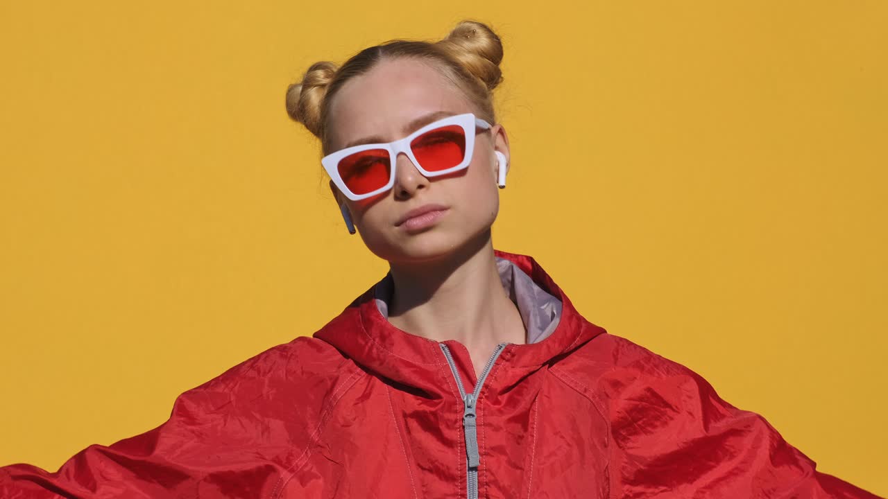 Portrait of happy teen blonde girl in airpods in pink glasses listens to music on smartphone, smiles, dances, shaking her head rhythmically against pink background of slow motion in summer. Emotions