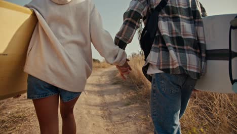 A-blond-guy-in-a-checkered-shirt-walks-hand-in-hand-with-his-blonde-girlfriend-in-a-white-sweatshirt-and-they-hold-surfboards-in-their-hands-and-walk-along-a-path-near-dried-grass-and-green-trees