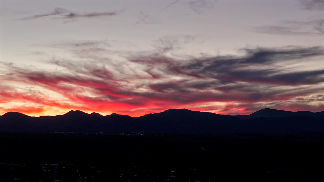 4K UHD drone timelapse capturing a vibrant, fiery red and orange sunset. Dramatic clouds move rapidly above the dark silhouette of a rugged mountain range while faint car lights trail below.