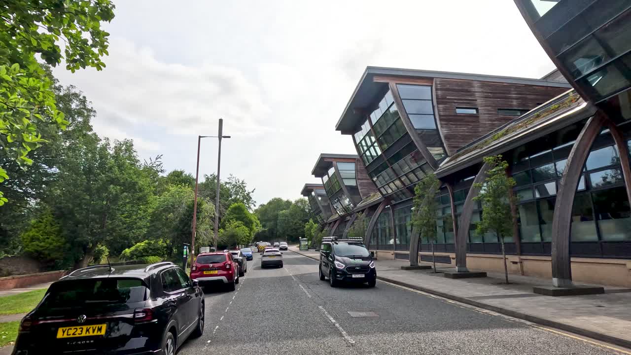 Cars travel along a tree-lined street beside a contemporary glass and wood academic building under daylight, with smooth camera motion suggesting a moving vehicle perspective