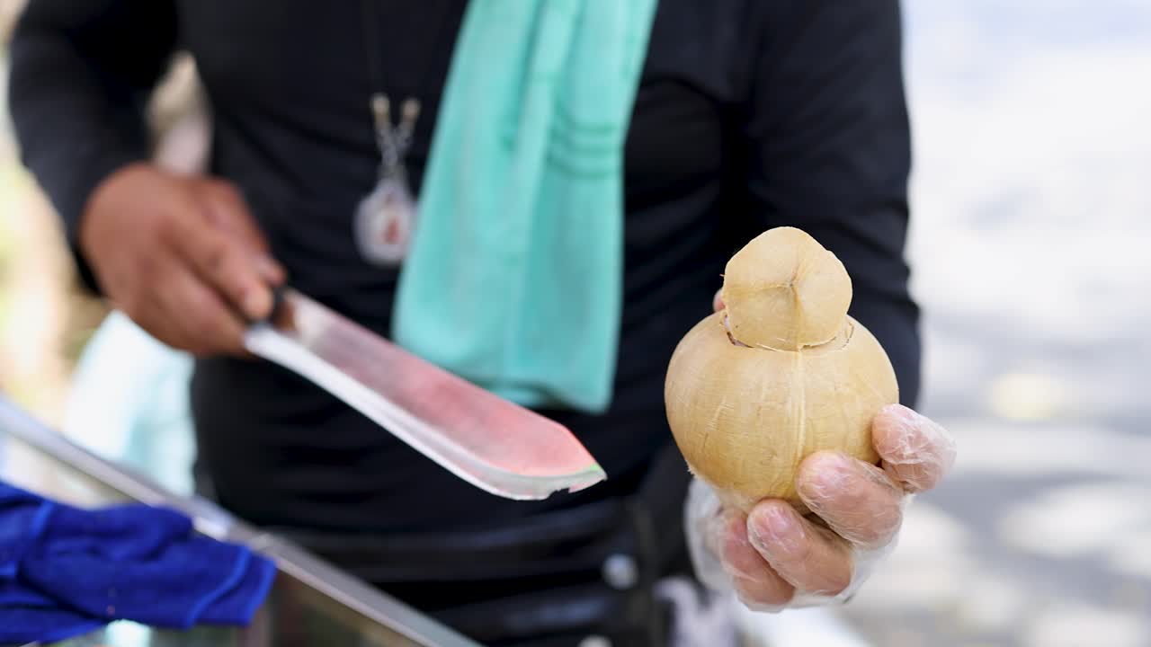 Vendor skillfully slices coconut with machete, wearing gloves, outdoors in bright natural daylight