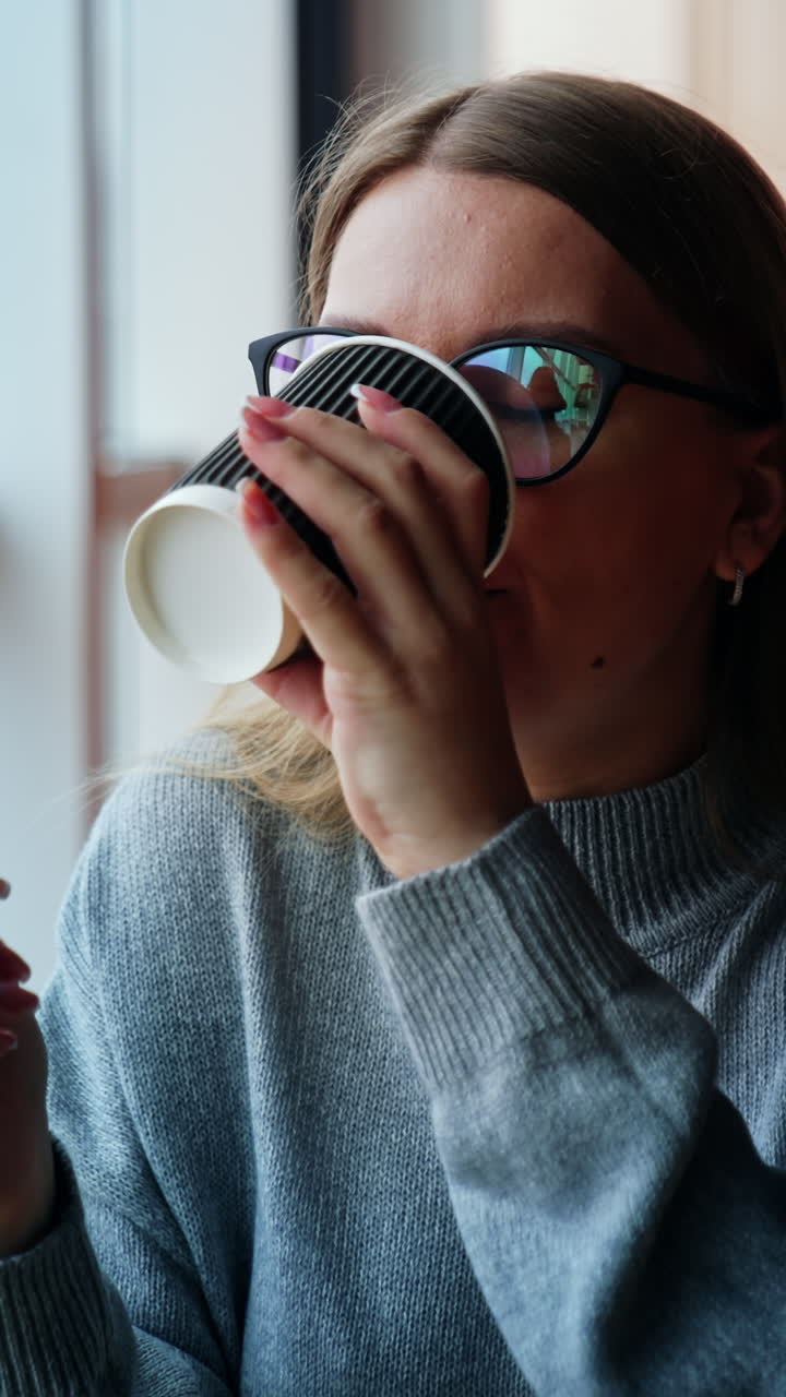 Smiling dreamy lady drinks coffee looking into window. Woman having a break from work at laptop. Lunch time at the working place. Close up. Vertical video