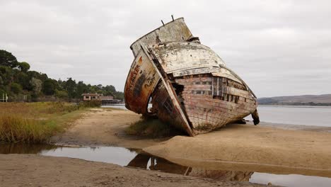 Panning-shot-of-rotting,-shipwrecked-Point-Reyes-boat-washed-up-on-a-cloudy,-marshy-Tomales-Bay-shore