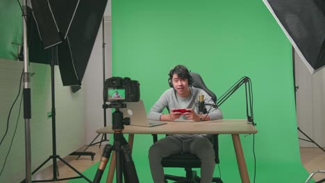 una cámara grabando a un hombre asiático sonriente jugando con auriculares y computadora mirando a la cámara mientras juega un juego de teléfono móvil en la pantalla verde con equipo de luz profesional
