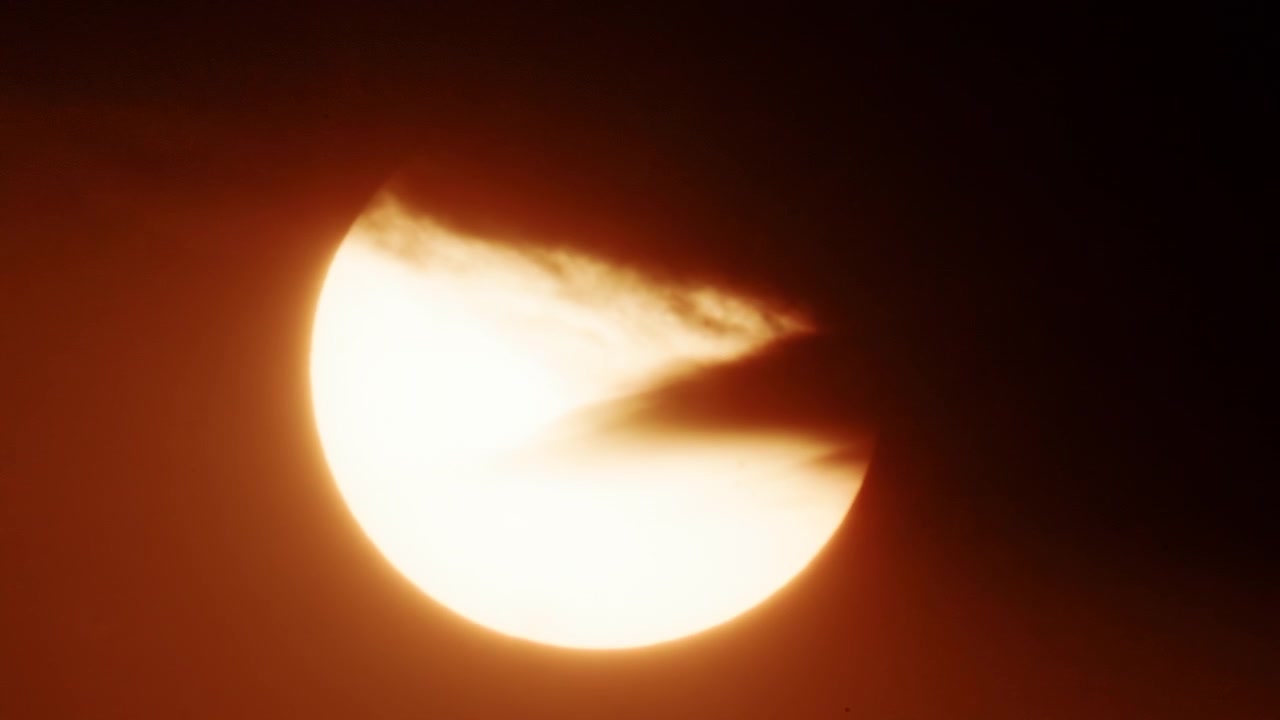 Close up of a bright sun partially covered by thick moving clouds, glowing in intense golden orange sunset tones