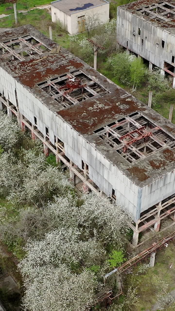 Destroyed factory buildings with holes in roofs. Old abandoned industrial plant. Ruined manufacturing on green nature background. View from above. Vertical video