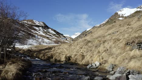 Luftaufnahme-Einer-Berglandschaft-In-Den-Pyrenäen-–-Tieffliegende-Drohnenaufnahme-über-Einem-Bach,-Ein-Tal-Hinauf-Und-An-Einem-Schutzhüttengebäude-Im-Winter-Vorbei