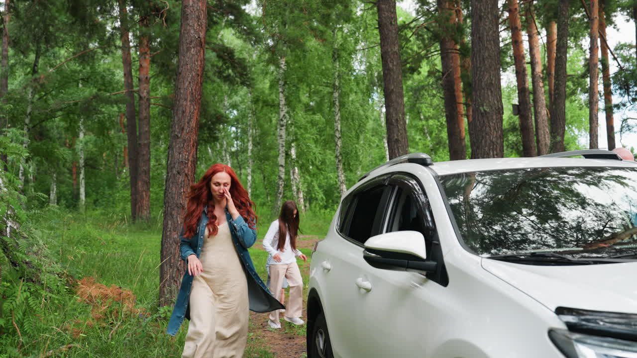 Stylish woman with red hair in long jacket and gown walks toward white car parked in forest while her daughters follow behind, bright summer greenery surrounds scene, calm family moment