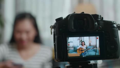 close up de un monitor de cámara grabando a una mujer asiática leyendo comentarios en su teléfono inteligente y hablando mientras transmite en vivo tocando la guitarra en una computadora portátil en casa