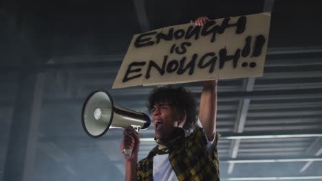 african american man wearing face mask talking in megaphone holding protest placard in empty parking