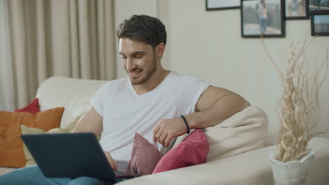 smiling man looking at laptop computer at home sofa. pensive guy working online