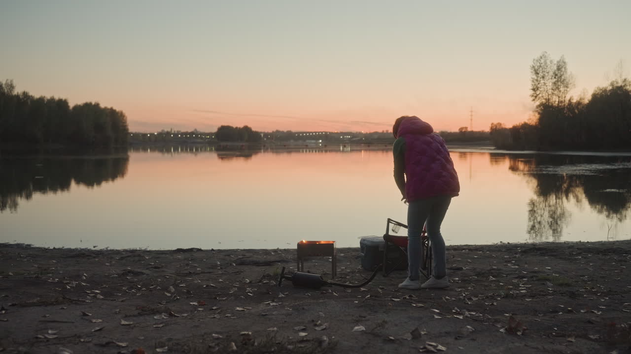 Camping lakeside sunset person preparing stove. Camper in warm jacket bends over portable stove on pebble shore, boiling kettle and arranging gear for meal, calm water reflection mirrors pastel sky