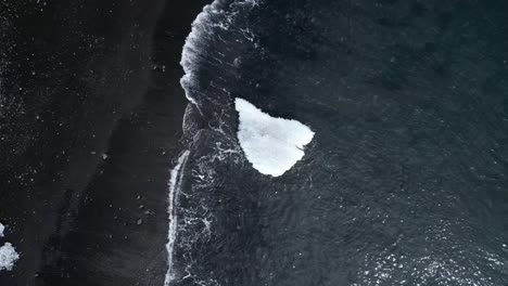 Playa-De-Diamantes-Desde-Una-Vista-Aérea-Centrándose-En-Las-Olas-Del-Mar-Moviéndose-Hacia-Un-Enorme-Iceberg-En-Las-Hermosas-Arenas-Negras-De-Este-Paisaje