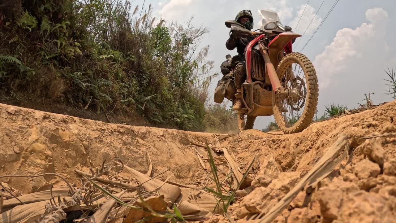 Terreno collinare arido con sentiero roccioso che si snoda tra alberi sparsi e copertura del terreno dorata, vista dal basso di un motociclista