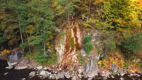 Forward-aerial-shot-of-a-small-waterfall-flowing-down-a-rocky-cliff,-surrounded-by-autumn-trees