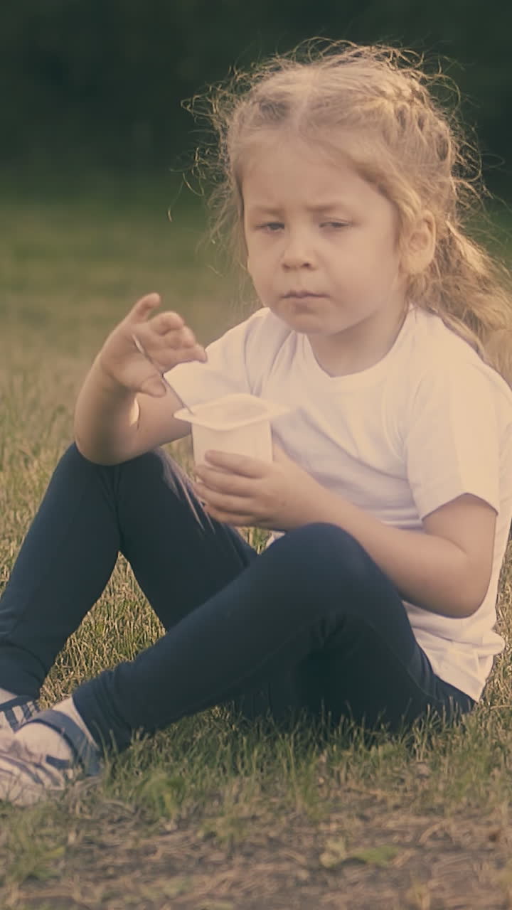 adorable little blond girl in white t-shirt eats tasty yogurt sitting on lawn grass in sunny park slow motion
