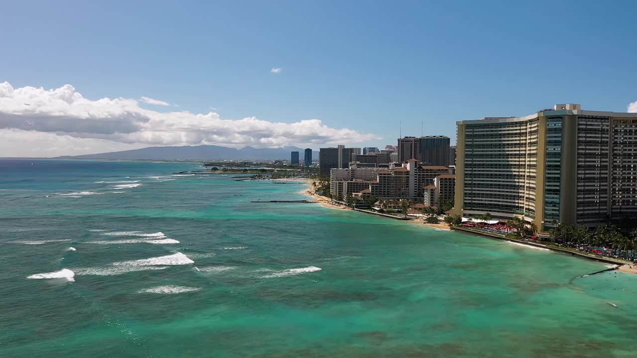 Aerial drone view showing Honolulu skyline with tall beachfront hotels, turquoise ocean water, and gentle waves along the coast of Oahu Island under bright blue sky in Hawaii