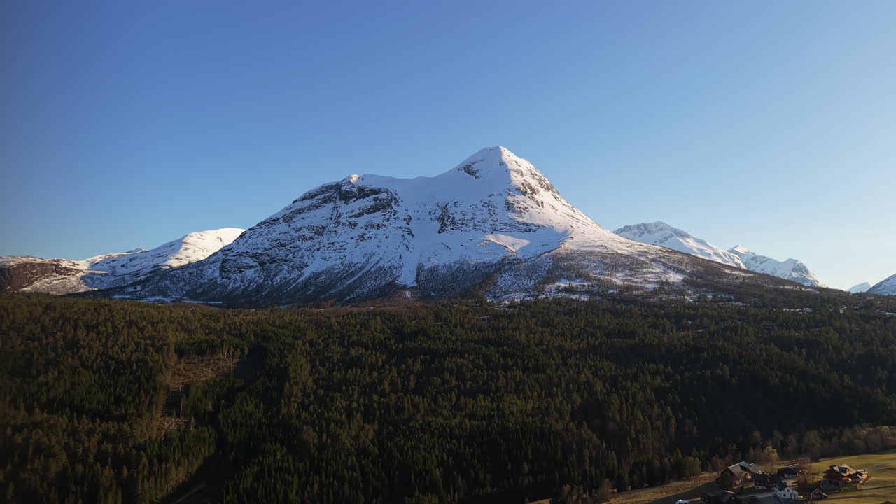Una maestosa montagna innevata si erge sopra una fitta foresta in Norvegia, sotto un cielo azzurro e limpido. Ideale per contenuti di viaggio o documentari naturalistici