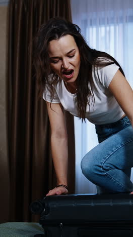 young woman struggling to close full suitcase. tired woman trying to close her travel luggage on bed in the hotel room. vertical video
