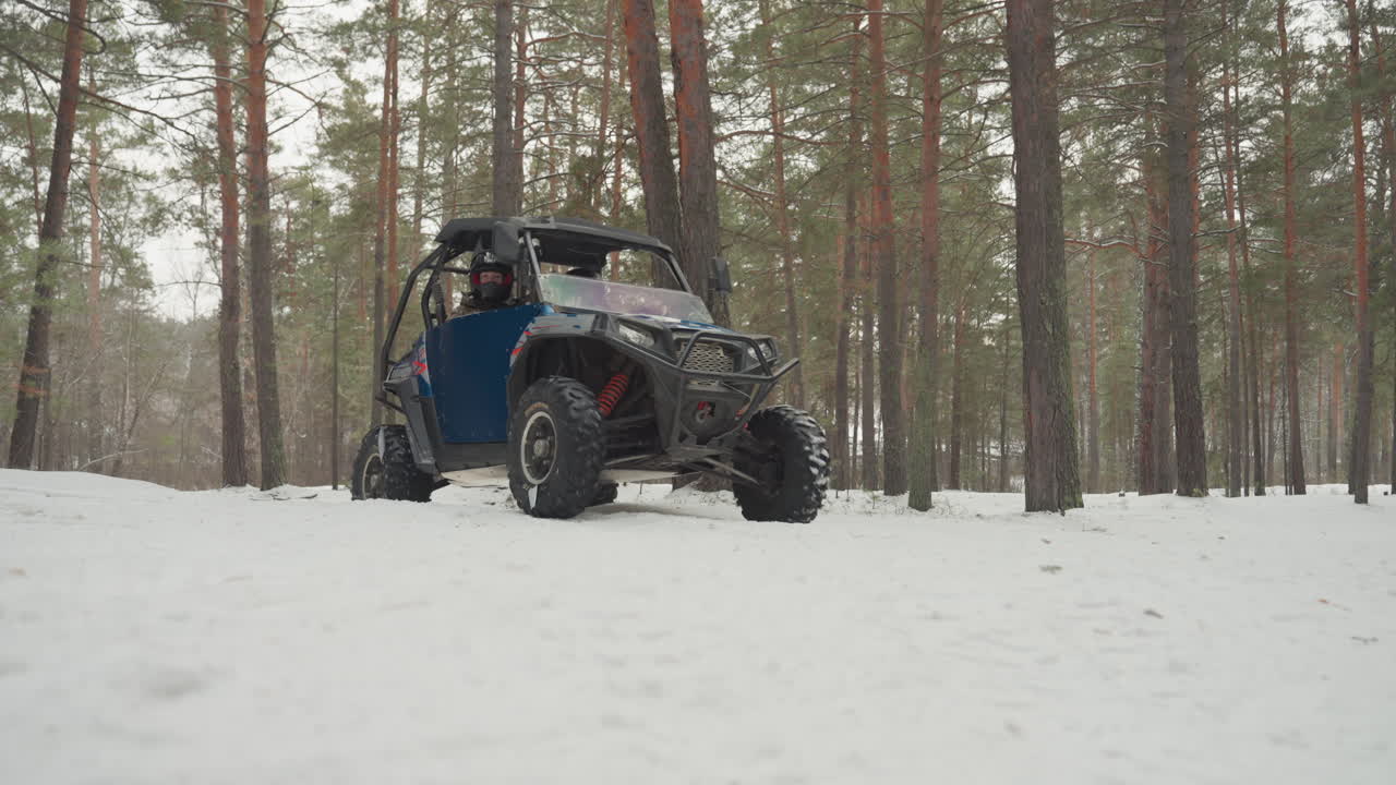 ATV blu che sfreccia attraverso una foresta di pini innevata, il pilota naviga tra i tronchi, raffiche di neve, primo piano da bassa angolazione di pneumatici robusti e tracce smosse, luce invernale coperta, atmosfera avventurosa e ad alta energia.