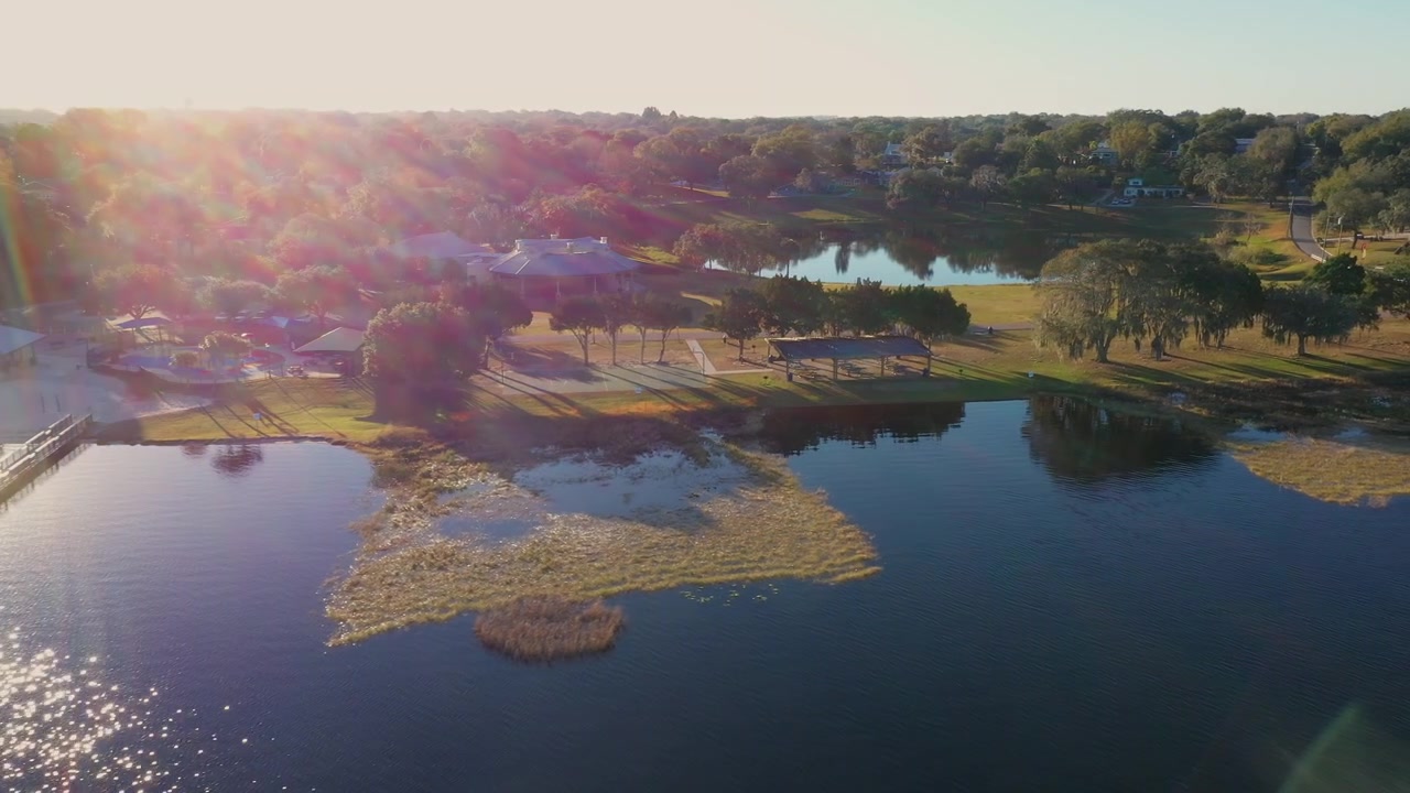Ripresa aerea con drone del molo e della spiaggia del Waterfront Park sul lago Minneola a Clermont, Florida, con padiglioni, alberi rigogliosi e acque blu calme che si estendono lontano all'orizzonte