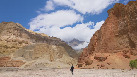 In-Upper-Mustang-Nepal,-A-tourist-walks-infront-of-Huge-muddy-rock-mountains-under-the-Blue-cloudy-sky