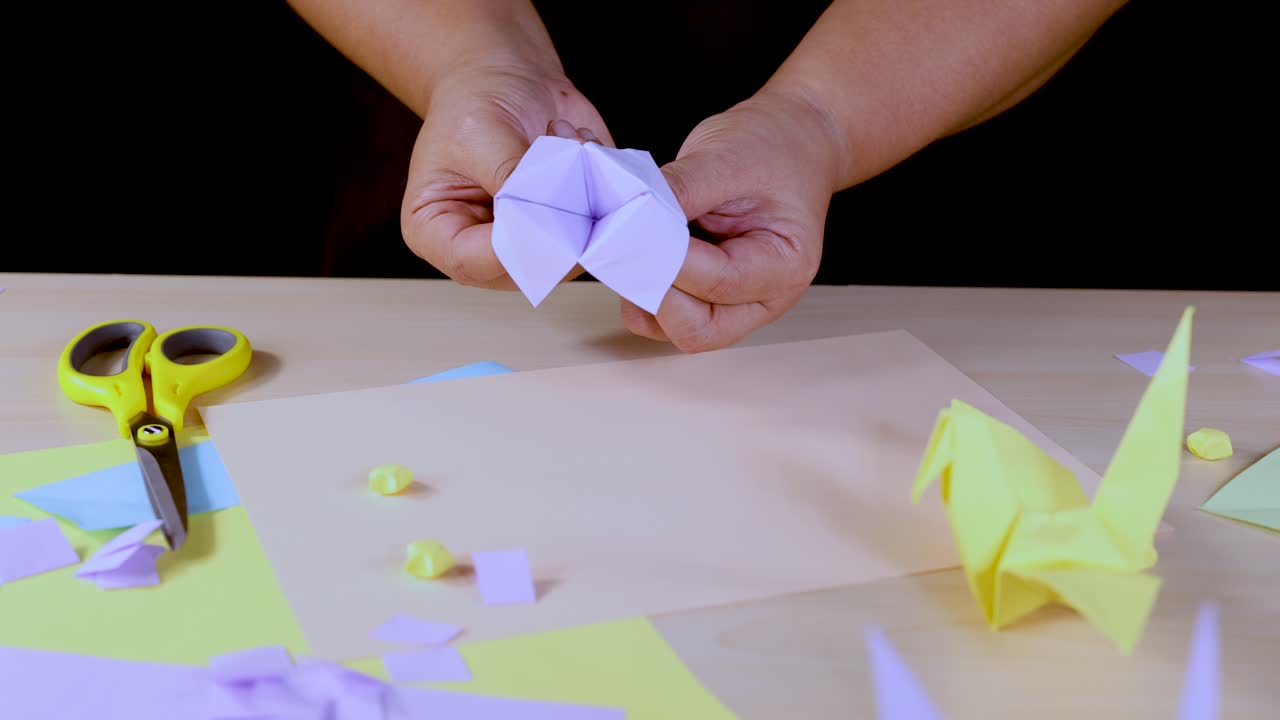 Person folds white origami fortune teller on table with scissors, paper, and yellow crane