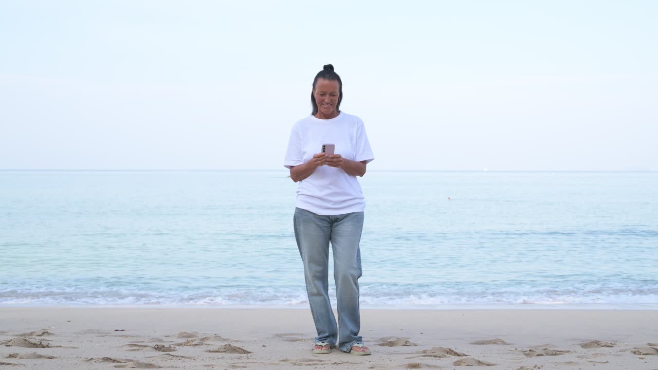 Skandinavisk moden turistkvinde iført 90'ernes normcore-tøj i Thailand, der bruger telefon på stranden