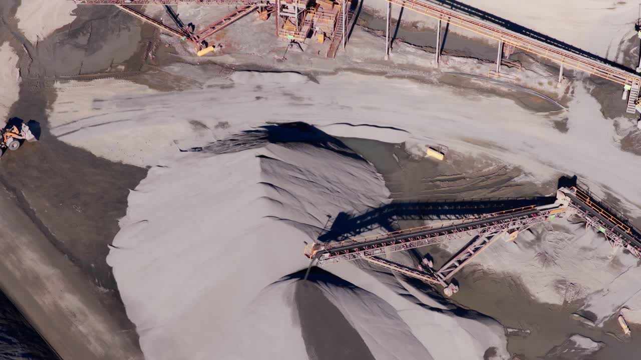 Top down aerial of conveyor belt casting shadow spreading over gravel pile as front loaders drive