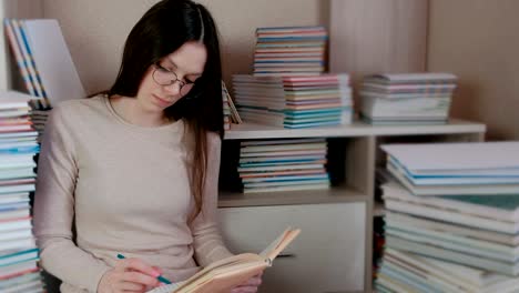 una joven morena con gafas escribe las notas sentada en el suelo entre los libros.