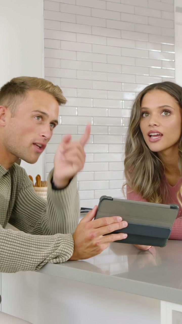 Vertikální video: Muž v zeleném svetru otáčí tabletem a sdílí obsah u pultu, žena v růžovém se usmívá.