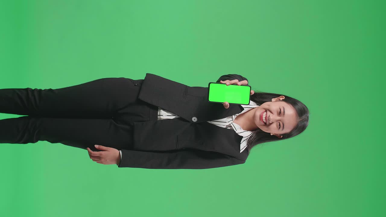 una mujer de negocios asiática con una sonrisa que muestra un teléfono móvil de pantalla verde mientras está de pie frente a la pantalla verde