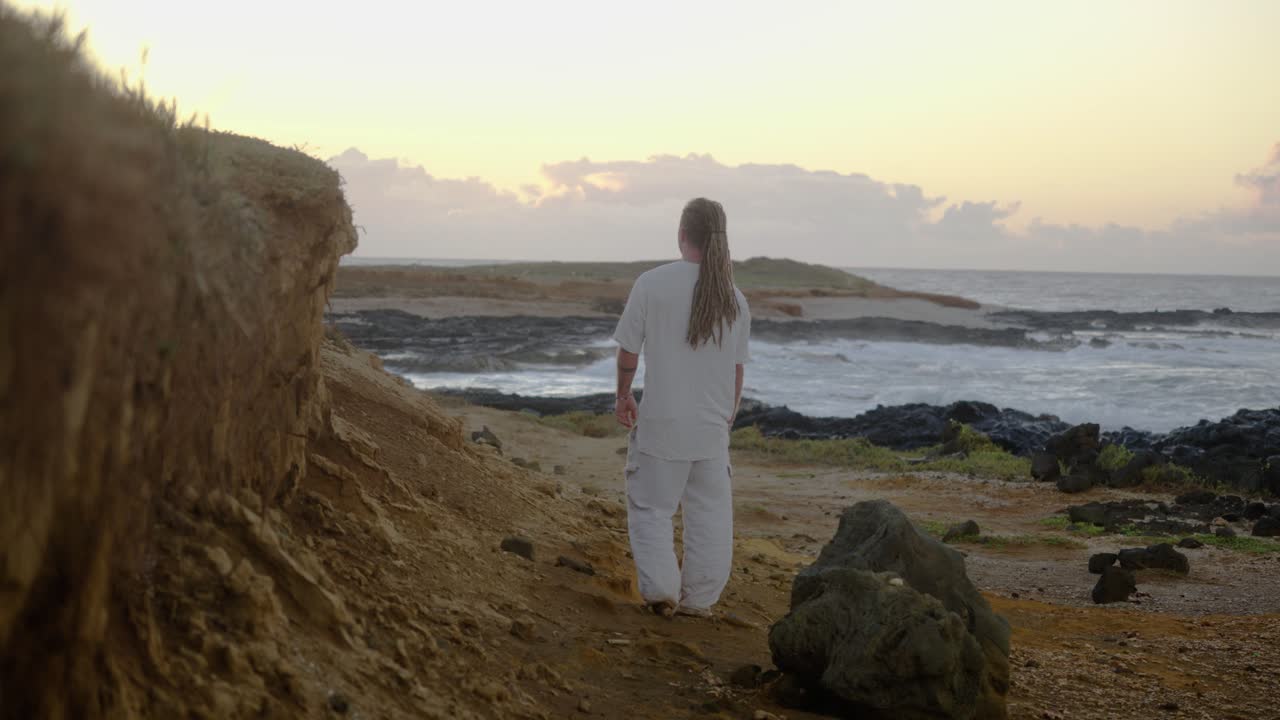 A man in white clothing walks away on a rocky coastal path, moving toward the distant ocean horizon as waves break against the shore at sunrise