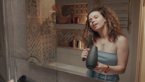 happy girl singing with hair dryer and comb in front of bathroom mirror