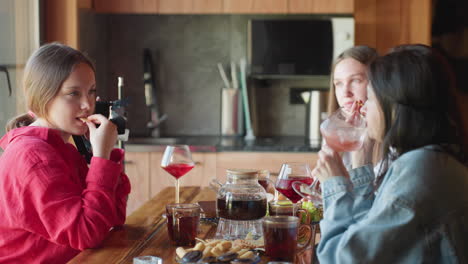 two women enjoy cozy moment together at wooden table with snacks and colorful drinks, one holding studio light playfully, warm natural kitchen light enhances relaxed friendly atmosphere