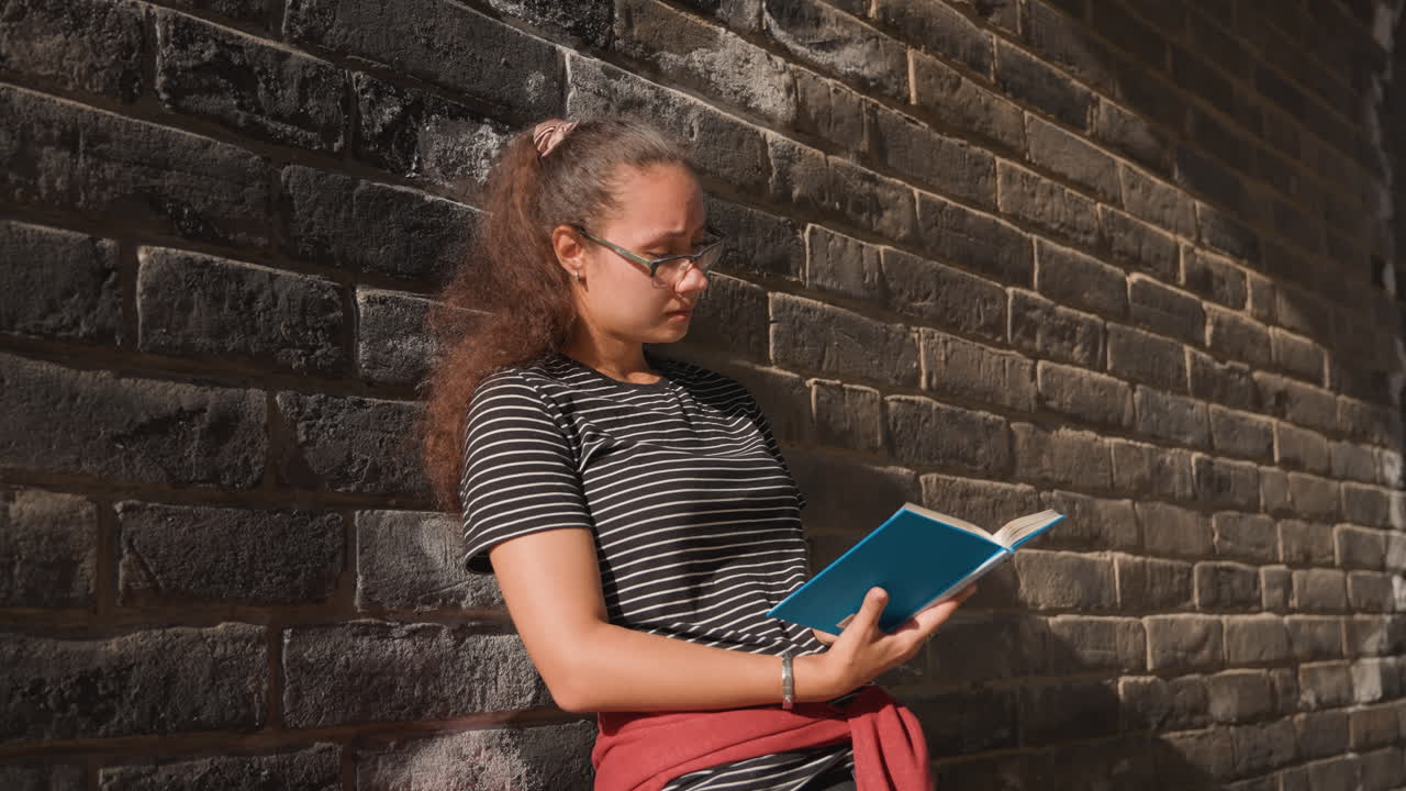 Jeune femme lisant contre un mur de briques, allée ensoleillée, chemise rayée et lunettes, pull rouge noué à la taille, livre de poche bleu ouvert sur les genoux, posture détendue et penchée, arrière-plan de ville diurne avec des éléments en pierre.