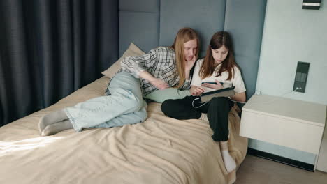 two teen girls using tablet and phone on a bed