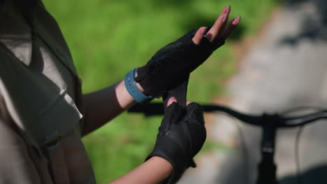 close-up back view of individual securing biker glove on left hand near bicycle, focusing on fingers adjusting strap with blurred background featuring lush greenery and paved pathway