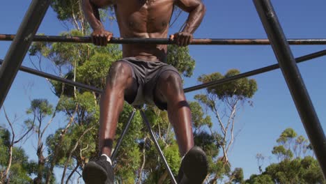 Fit, shirtless african american man exercising outside, doing hanging pull ups on a climbing frame