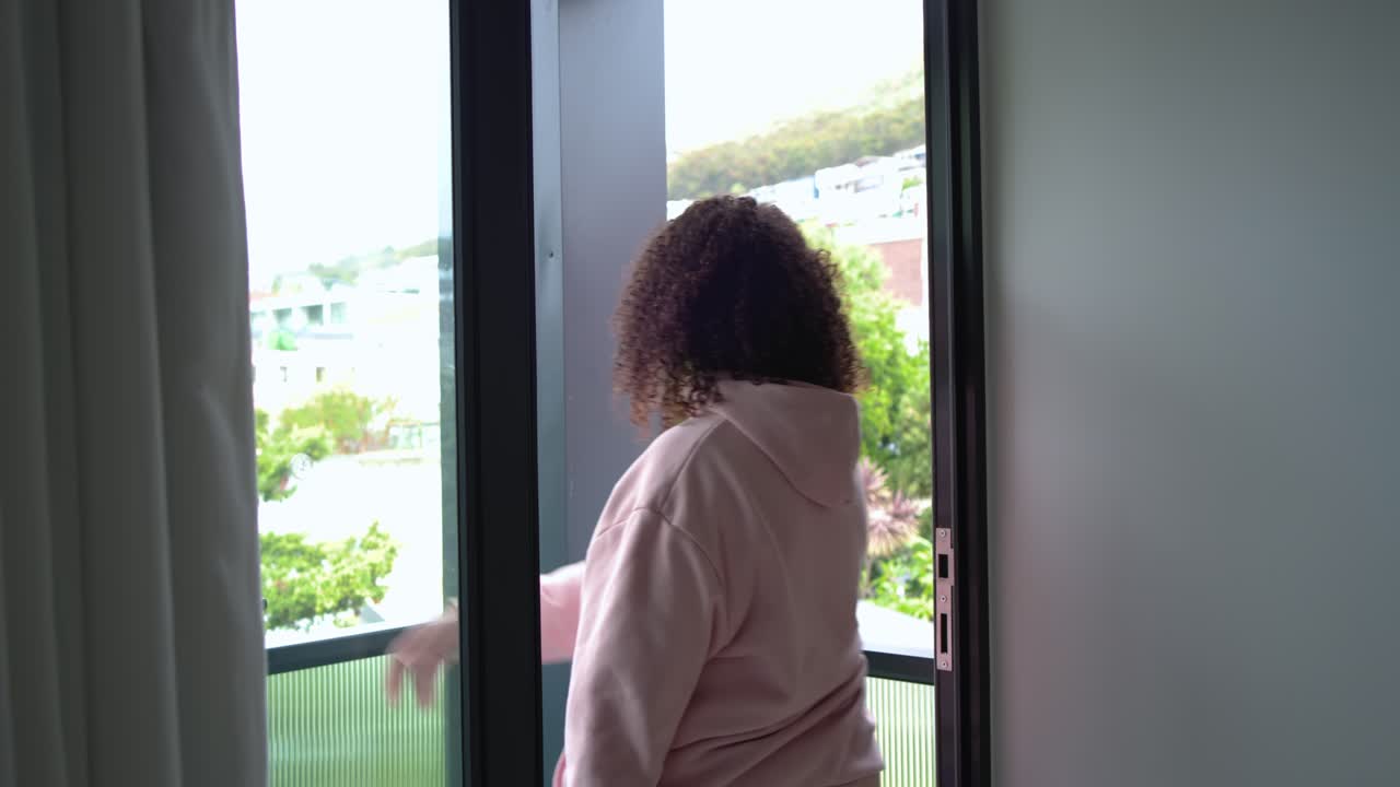 Pink-hooded woman is pulling curtain, opening sliding door, stepping onto balcony to breathe