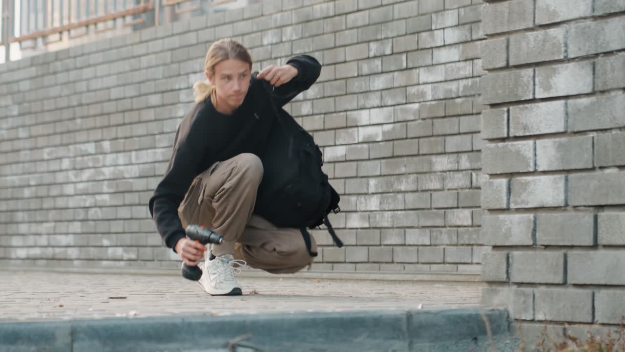 Uomo bianco accovacciato vicino a un muro di mattoni, che controlla lo zaino, ispeziona la cerniera vicino alle sneakers, con pantaloni cargo e maglione nero, su un marciapiede urbano, sotto una luce soffusa e cielo coperto, un'atmosfera da studente introspettivo, un momento di pausa.