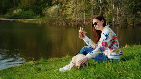 una mujer se sienta en un césped cerca de un lago, come comida rápida, al lado de un perro