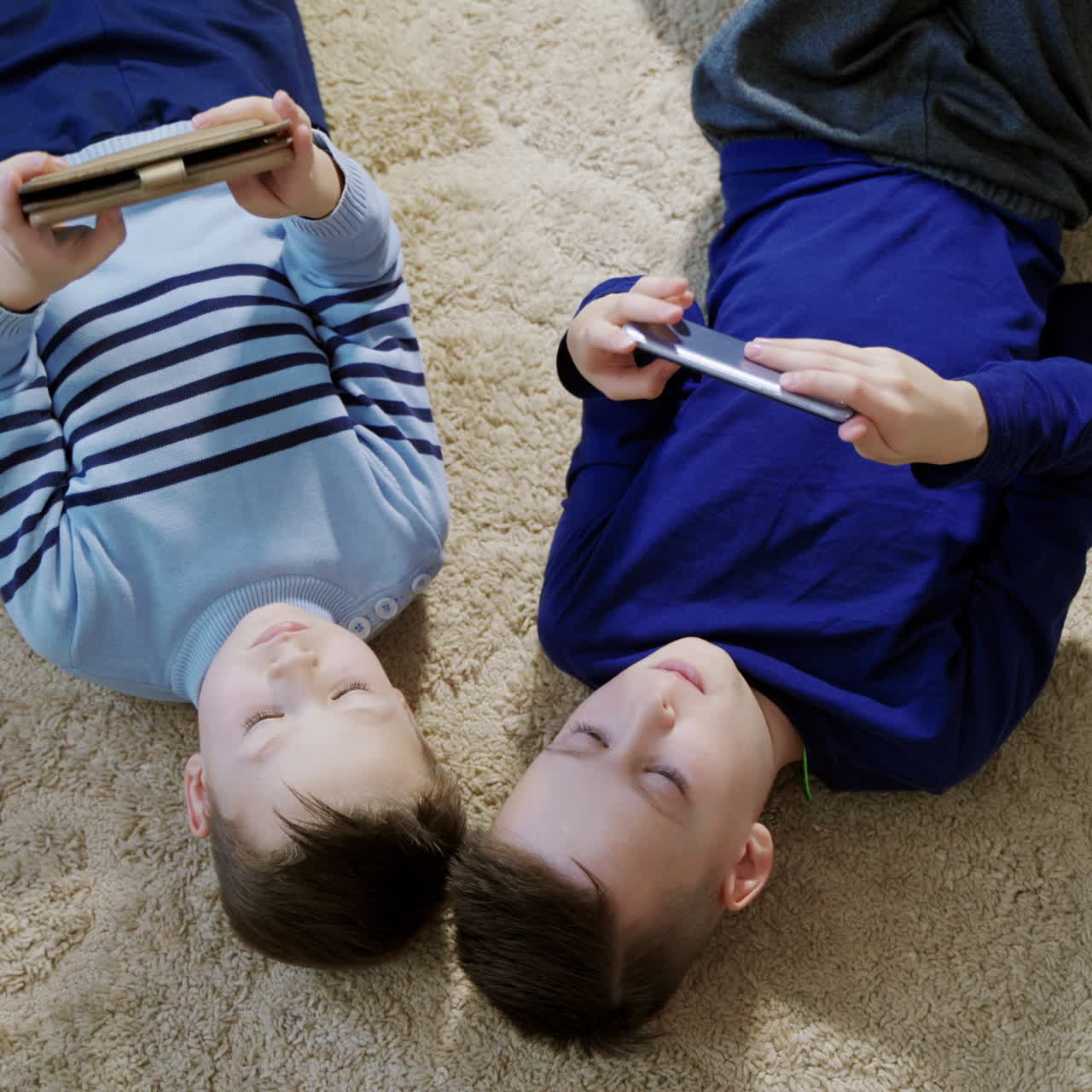 Boys lying on the floor on a light carpet and playing games on mobile phones at home. Top view of two kids playing together and helping each other in games.