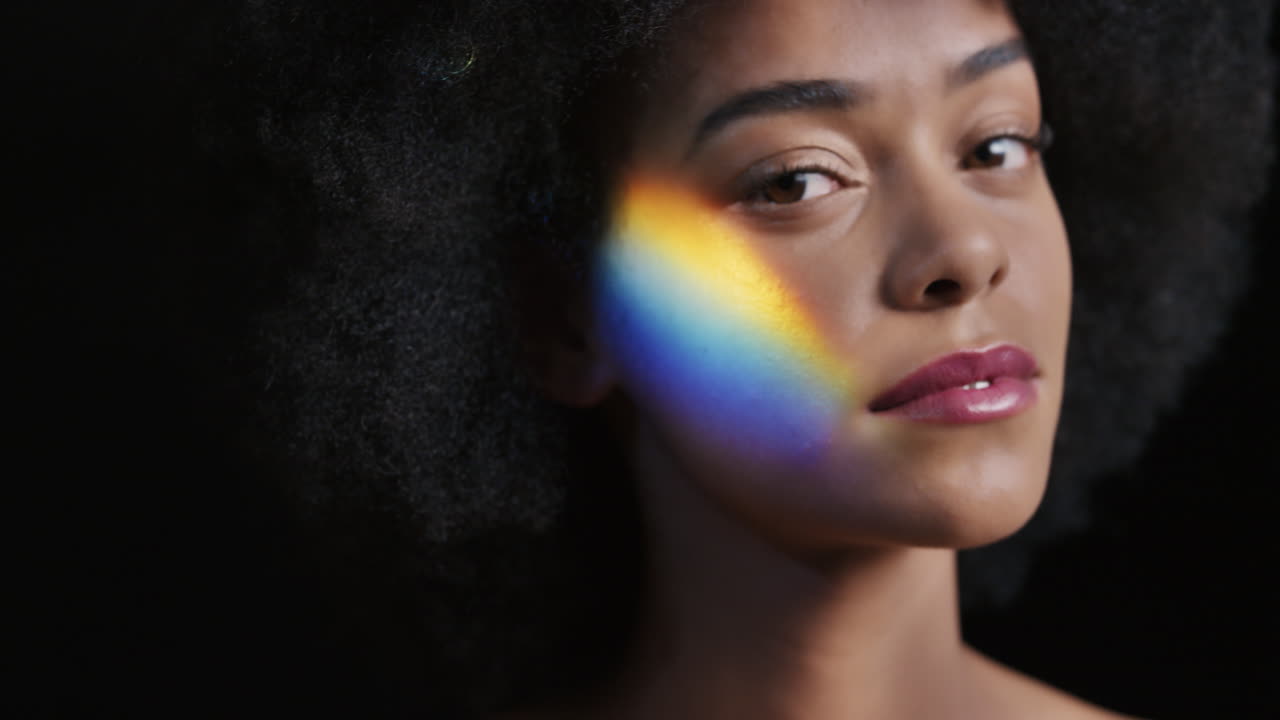 retrato de arco iris de cerca mujer afroamericana atractiva con afro disfrutando de una piel suave y saludable con aspecto confiado belleza natural luz multicolor sobre fondo negro concepto de cuidado de la piel